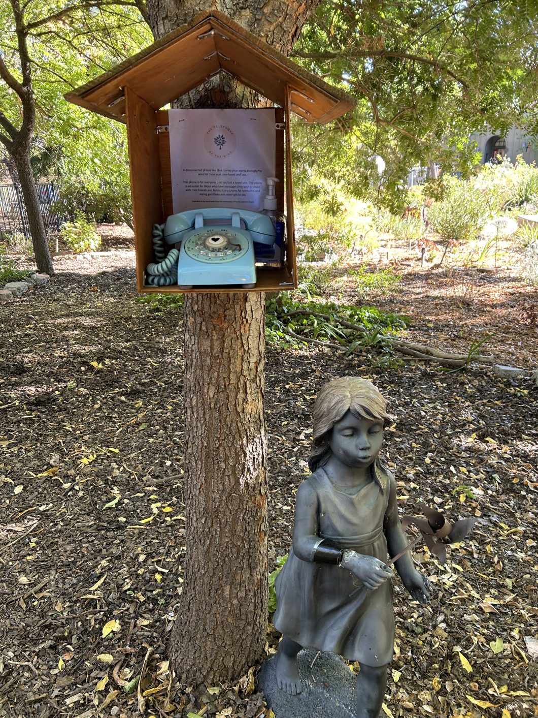 Of the Peace Wind Phone in the Peace Journey Forest, beyond the Labyrinth. A statue of a girl stands next to the phone. The blue rotatory phone has a wooden "house" around it on the trunk of a tree.
