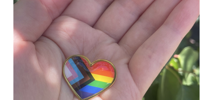 Pride Flag pin tenderly held in a person's hand. Background is the green leaves of a tree.