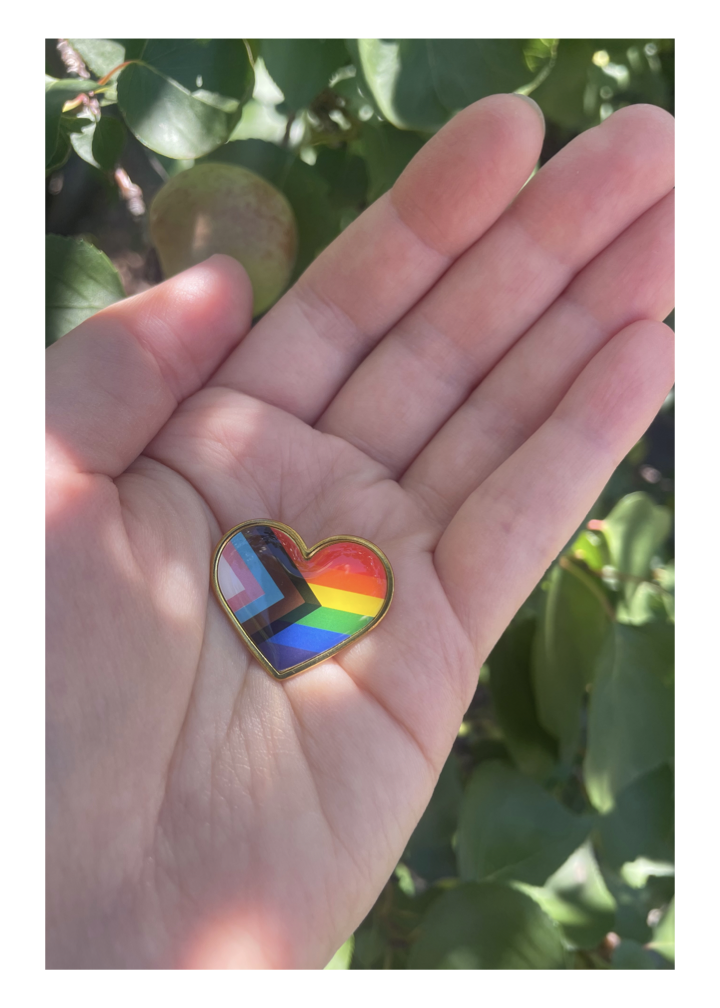 Pride Flag pin tenderly held in a person's hand. Background is the green leaves of a tree.