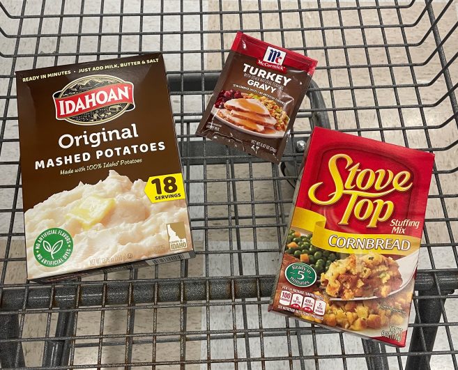 Box of instant mashed potatoes, gravy packet, and stuffing mix in a cart at a grocery store